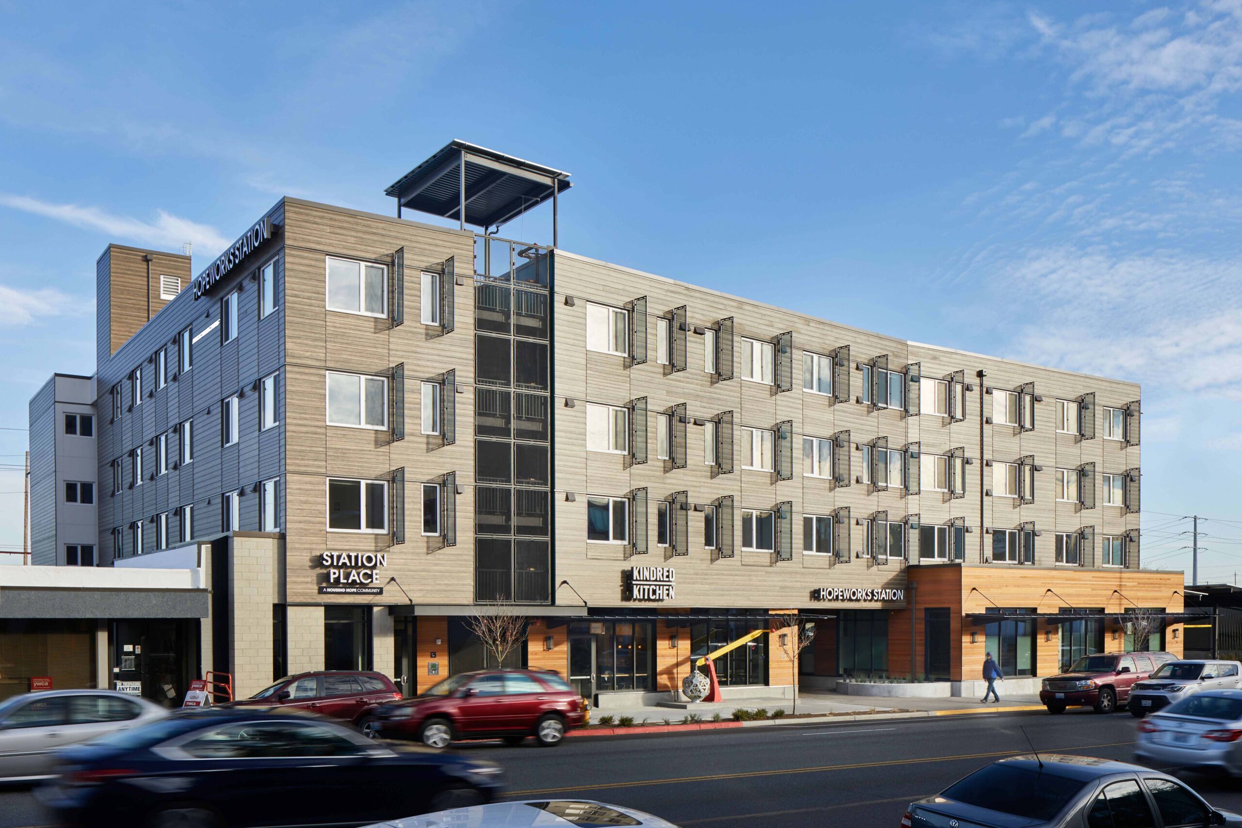 HopeWorks Station, a modern mixed-use building in Seattle, WA, featuring retail spaces on the ground floor and residential units above. The façade includes textured siding, operable window shades, and a rooftop terrace.