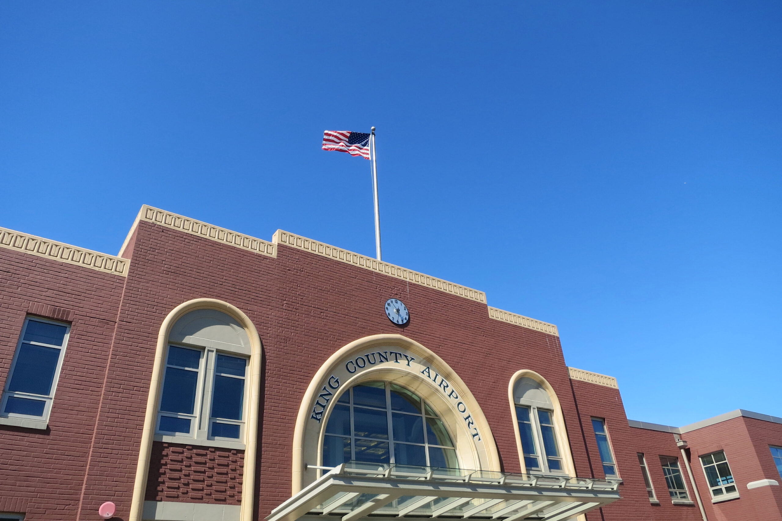 The main terminal building of King County International Airport (Boeing Field) in Seattle, WA, featuring a red brick façade, arched windows, a clock, and an American flag flying above against a clear blue sky.