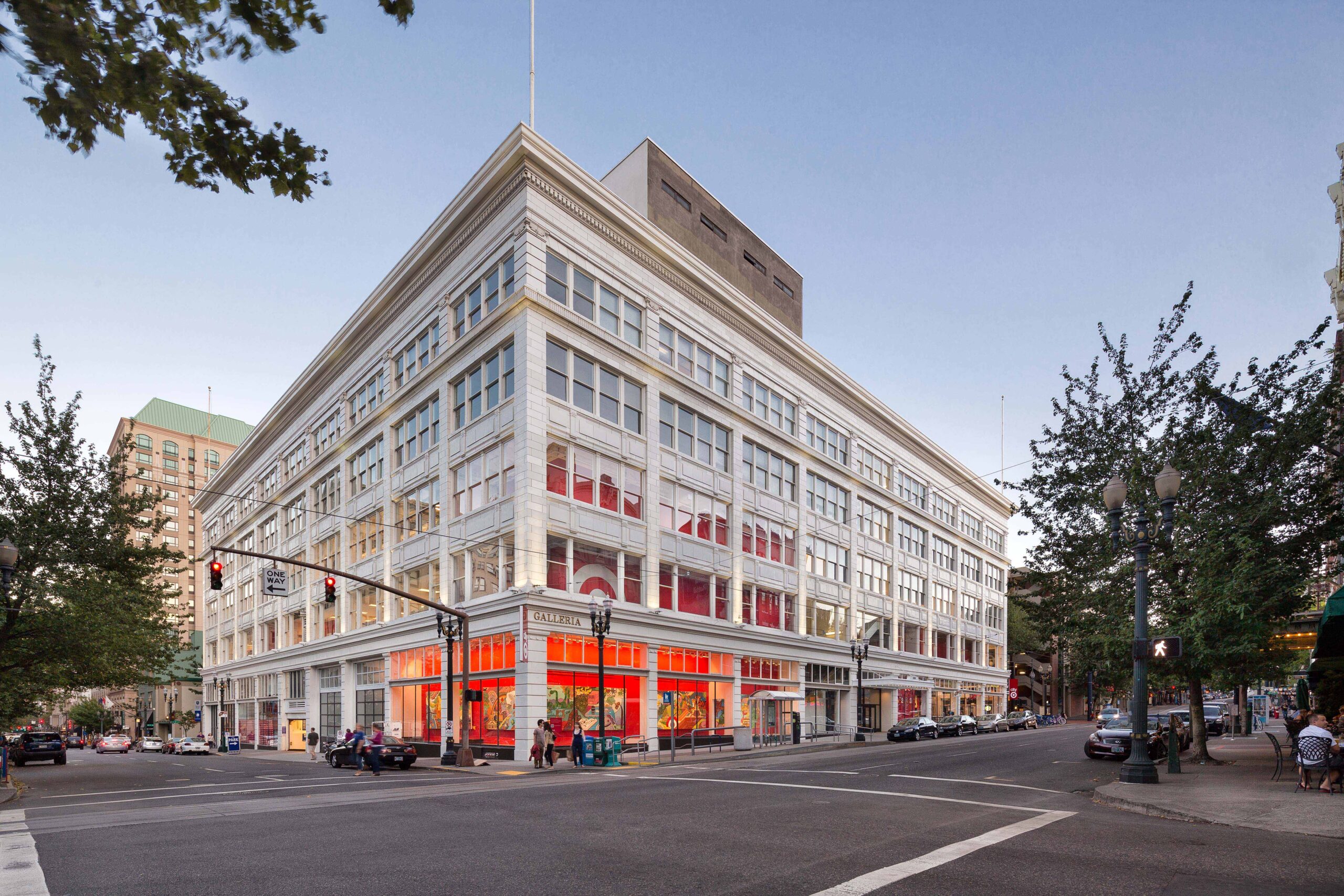 The Galleria Building in Portland, Oregon, a historic multi-story structure with a white façade and large windows. The corner of the building features a Target store with red signage and bright interior lighting visible from the street. The urban setting includes traffic lights, pedestrians, and surrounding buildings under a clear evening sky